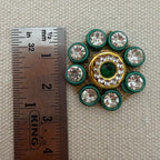 Decorative button with green and gold accents next to a ruler for scale on a textured beige background