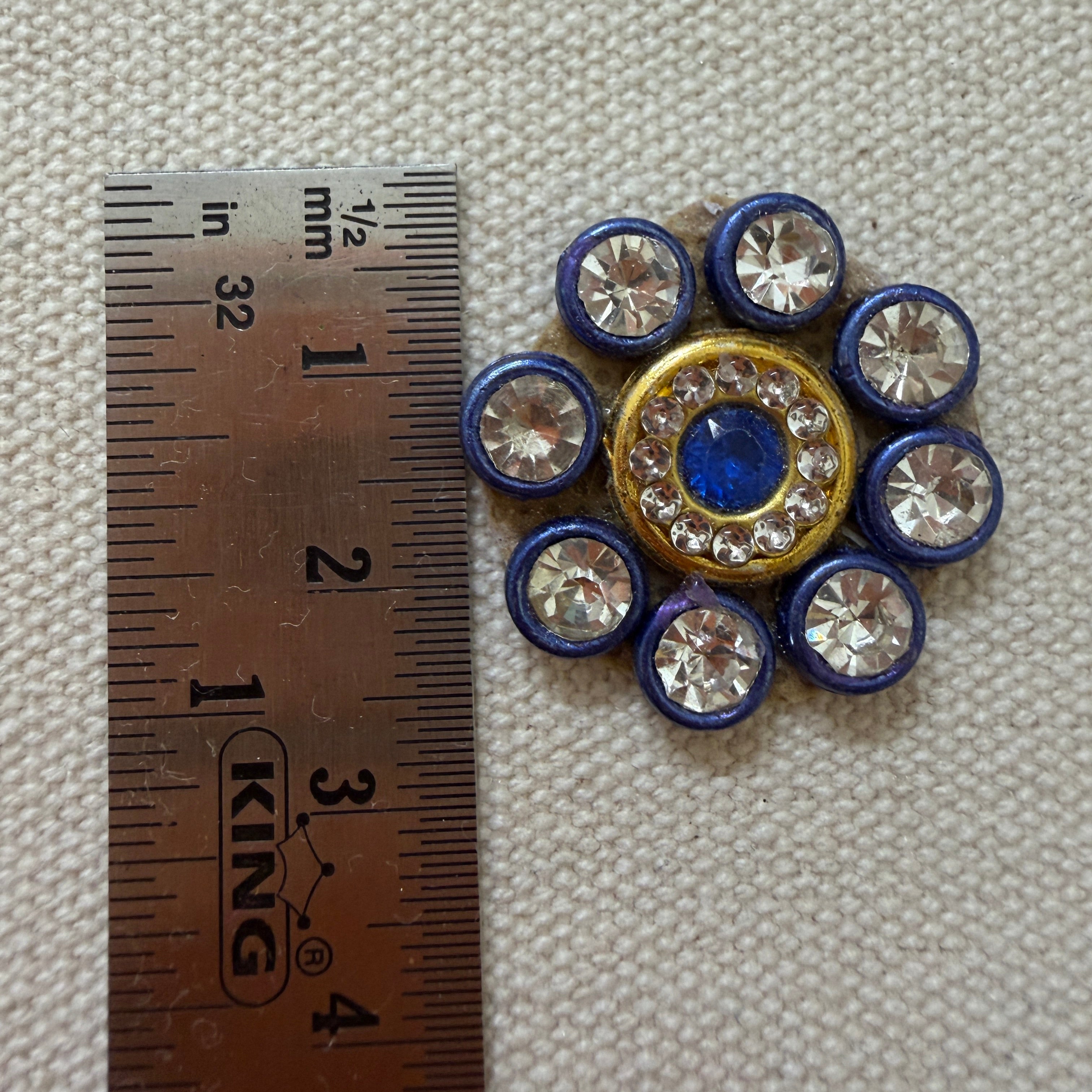Decorative button with blue and gold center and surrounding crystals, next to a ruler for scale.