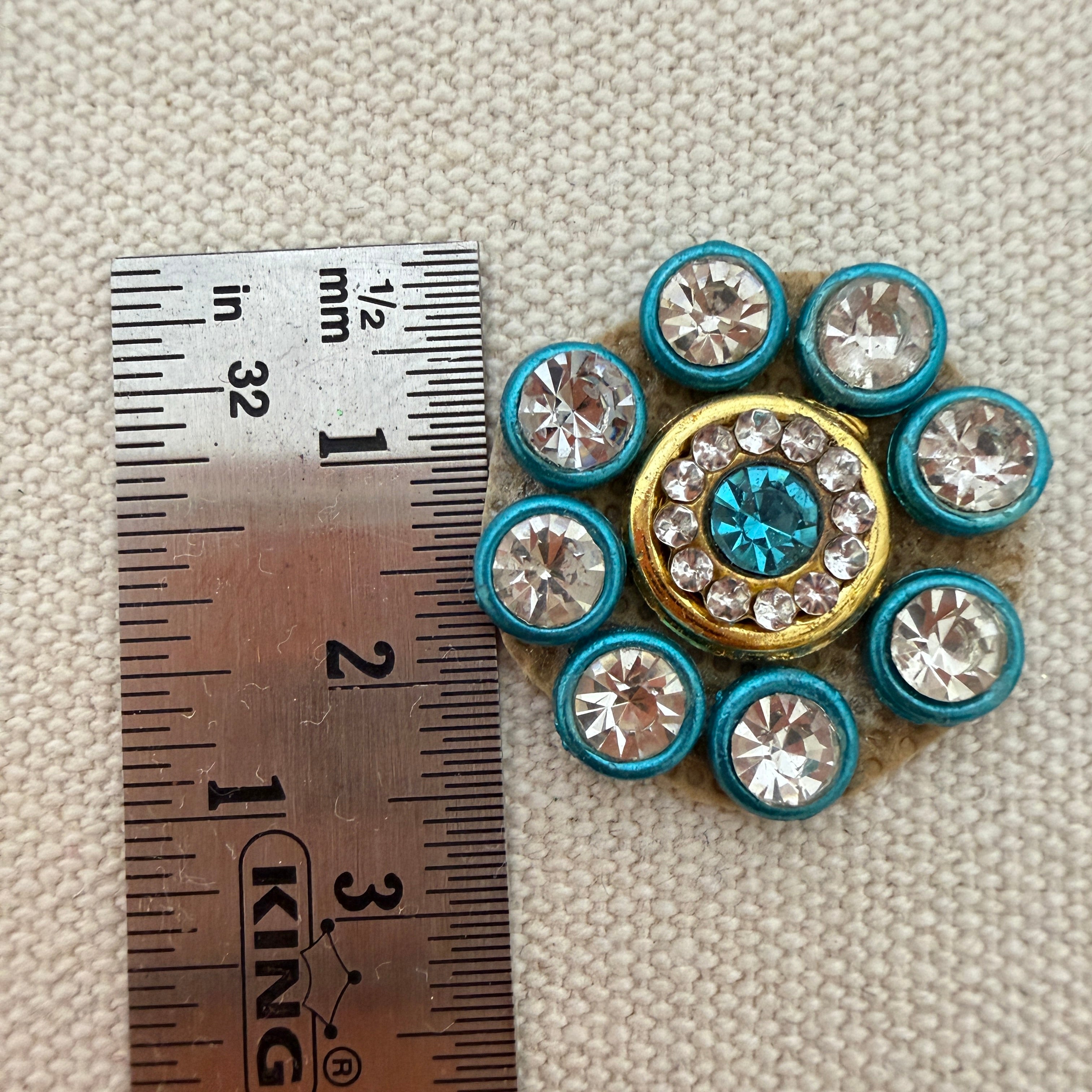 Decorative button with rhinestones next to a ruler for scale on a textured beige background