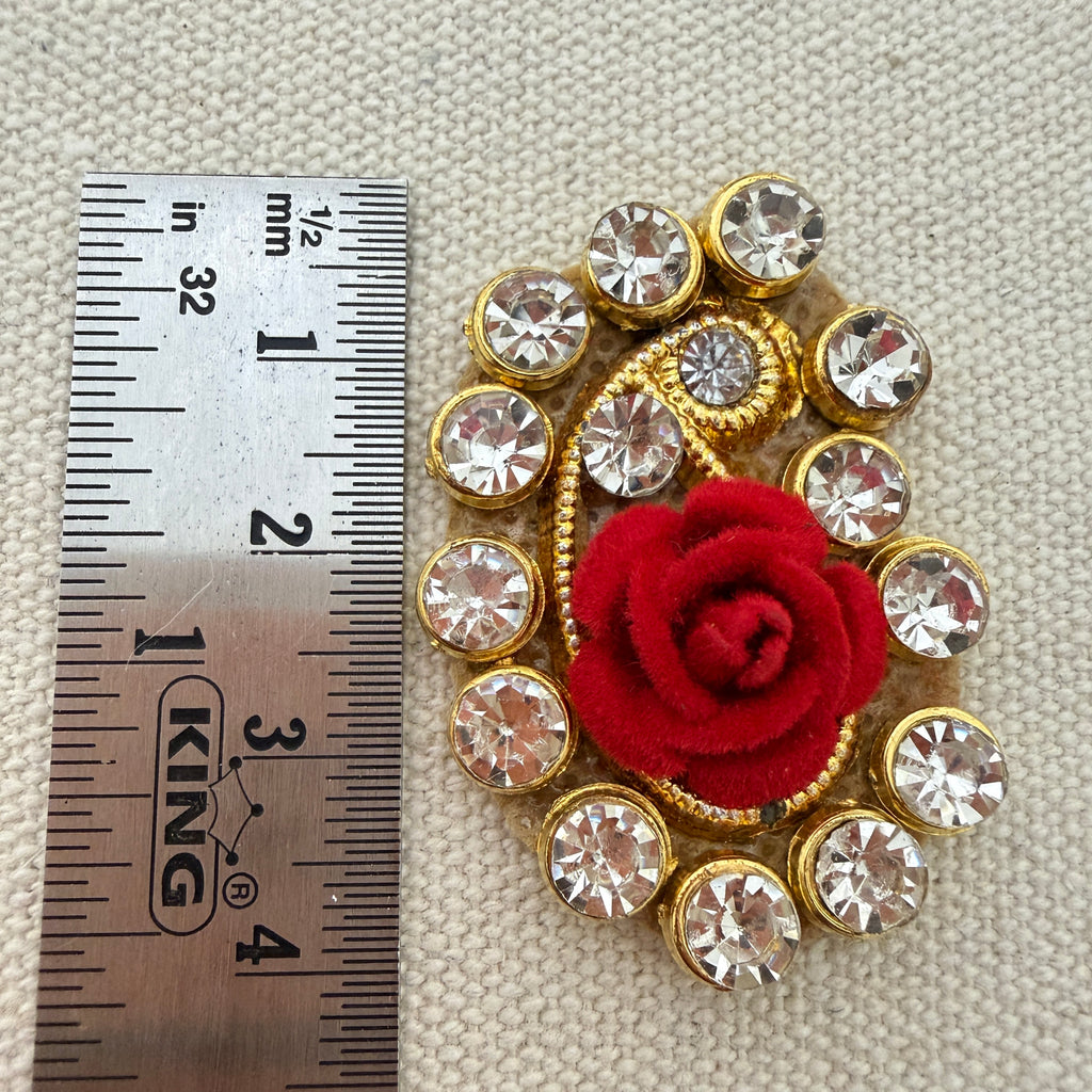 Decorative brooch with red flower and clear crystals next to a ruler for scale on a textured beige background