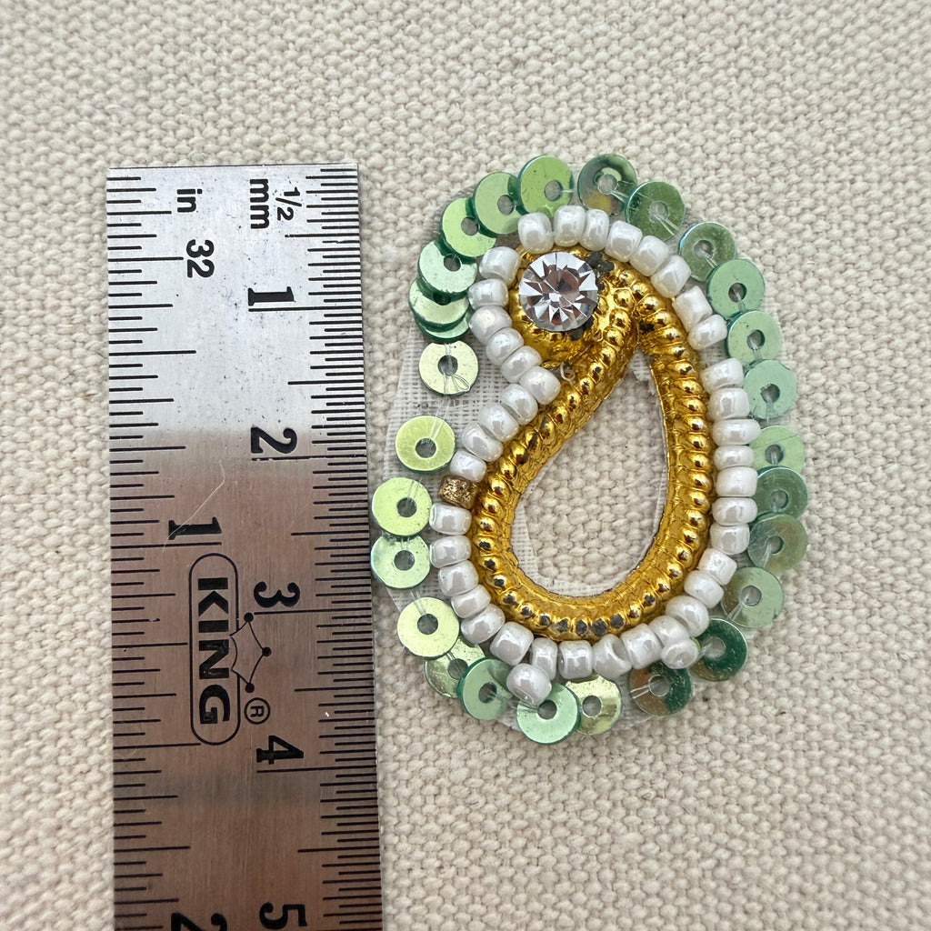 Decorative brooch with sequins and a crystal next to a ruler for scale on a textured beige background