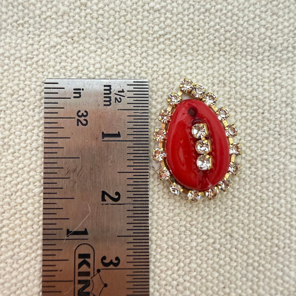 Red brooch with gold accents next to a ruler on a textured beige background