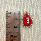 Red brooch with gold accents next to a ruler on a textured beige background