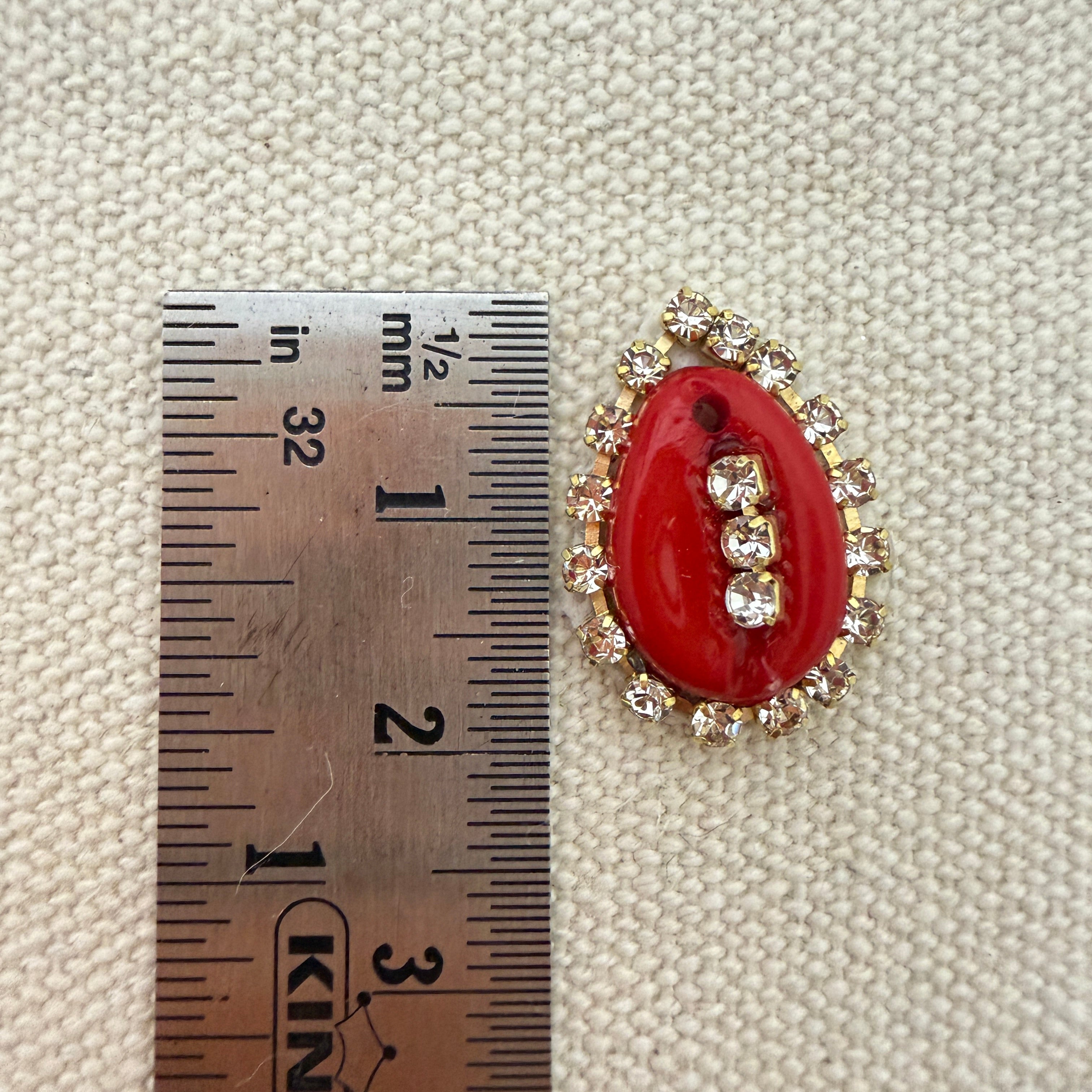 Red brooch with gold accents next to a ruler on a textured beige background