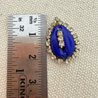 Blue brooch with gold accents next to a ruler on a textured surface