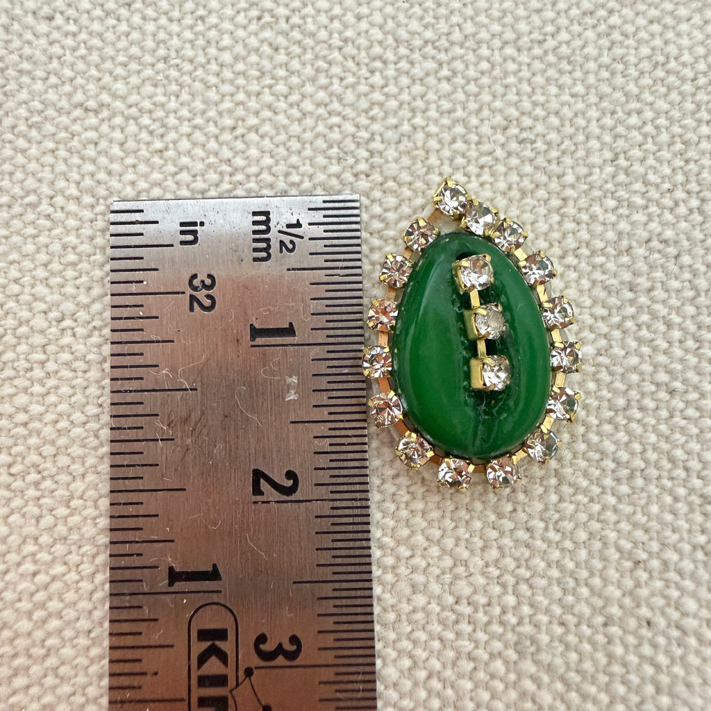 Jewelry item with green stone and gold accents next to a ruler for scale on a textured beige background