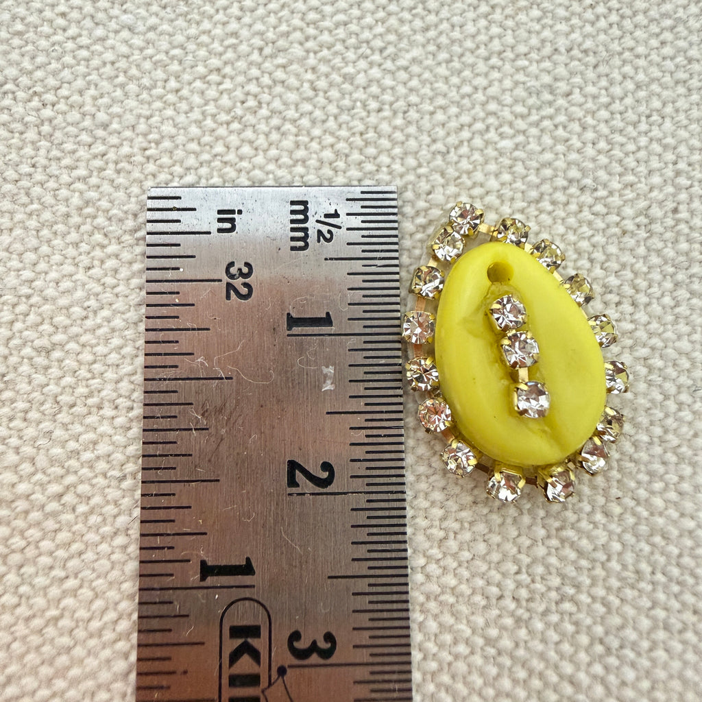 Yellow oval pendant with rhinestones on a textured beige background with a ruler for scale.