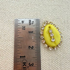 Yellow oval pendant with rhinestones on a textured beige background with a ruler for scale.