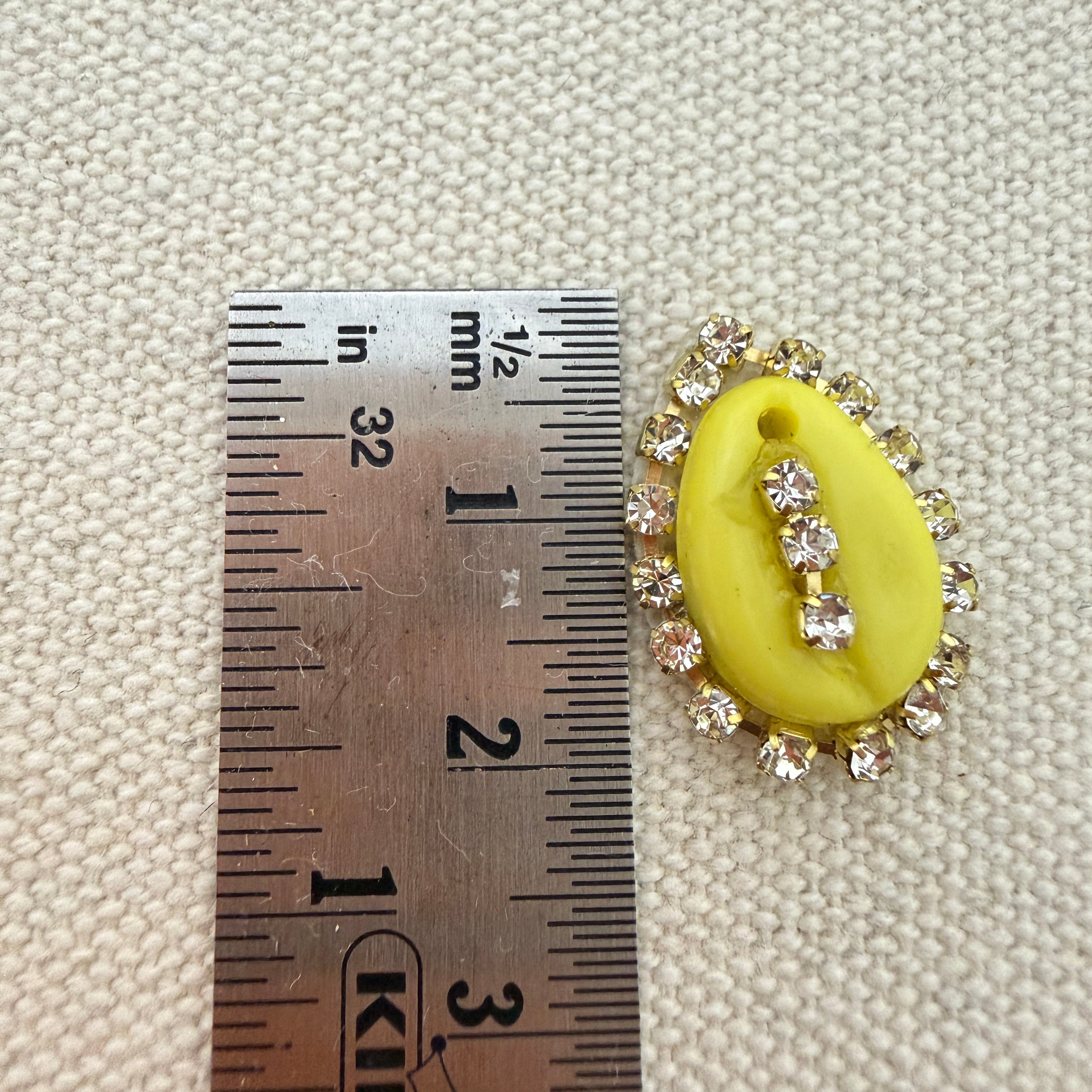Yellow oval pendant with rhinestones on a textured beige background with a ruler for scale.