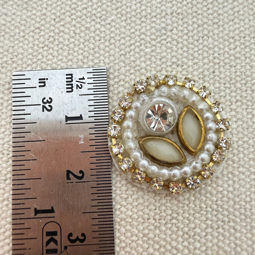 Decorative brooch with pearls and crystals next to a ruler on a textured beige background