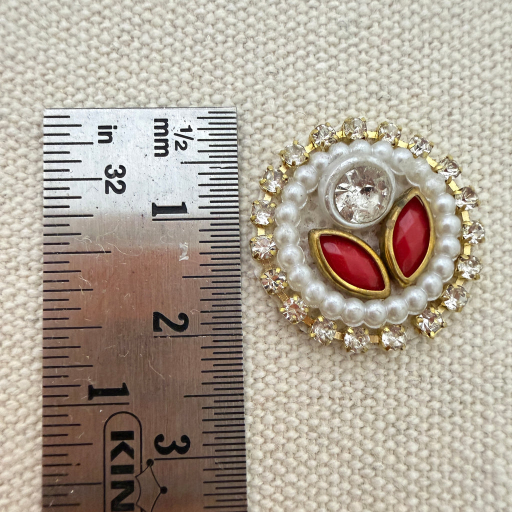 Jewelry piece with red stones and pearls next to a ruler for scale on a textured beige background