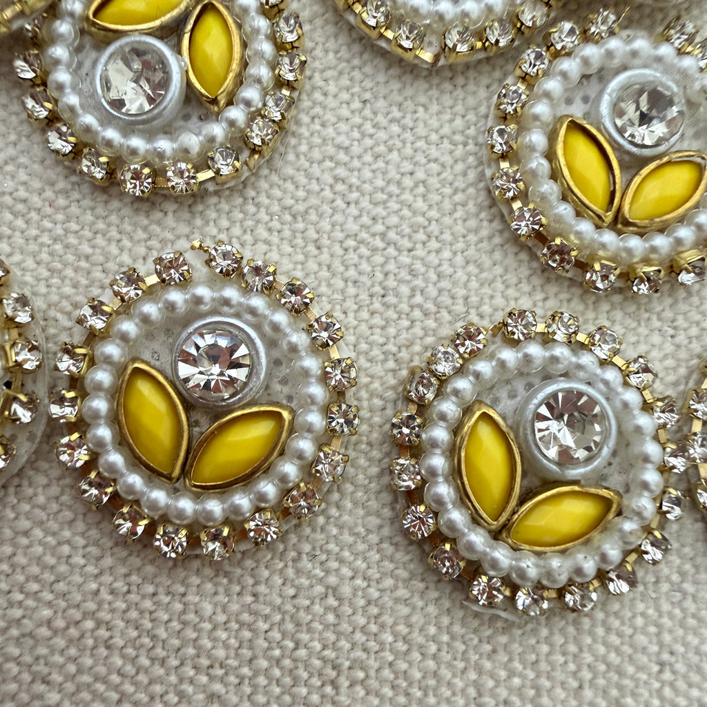 Decorative buttons with yellow center, pearl border, and crystal accents on a textured beige background.