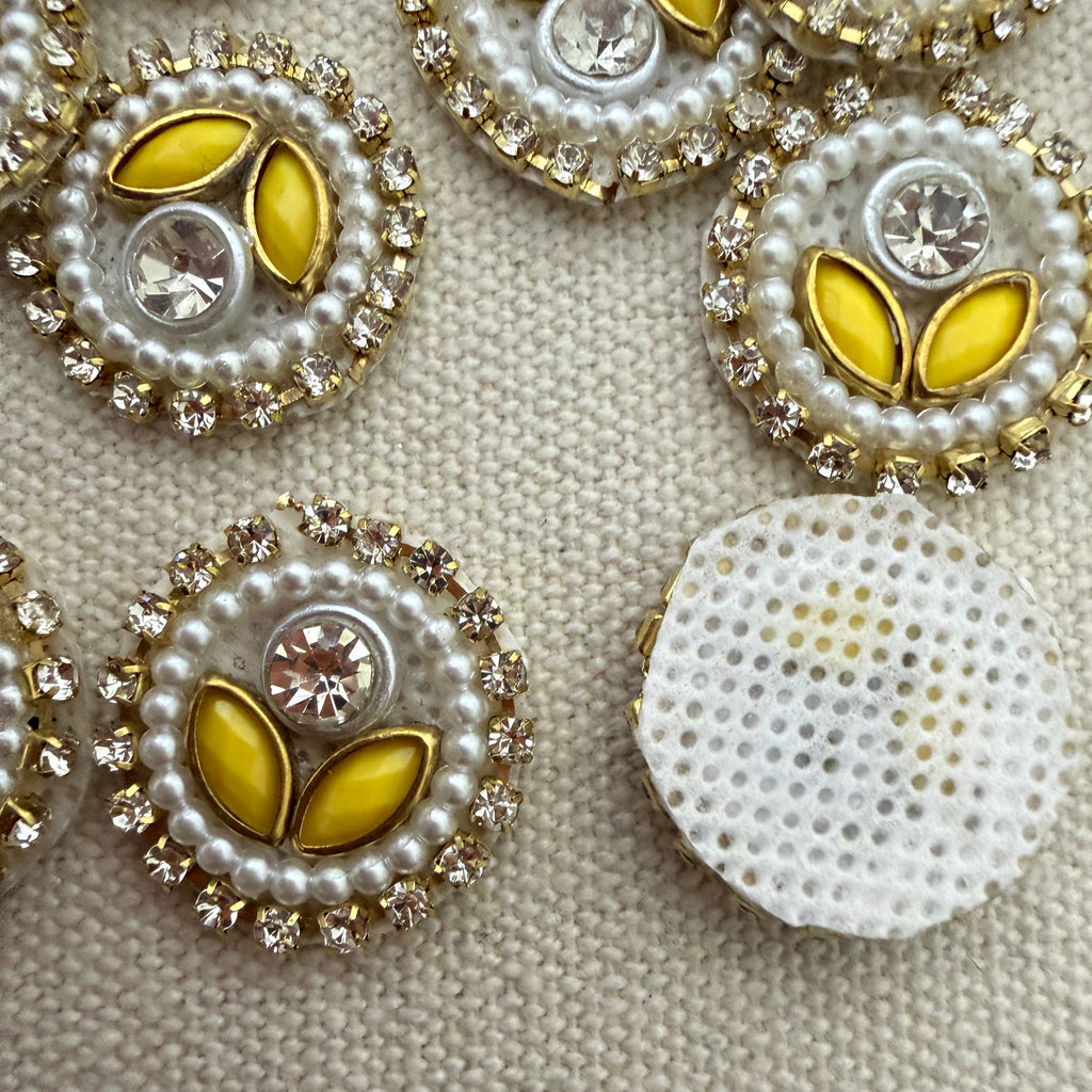 Close-up of decorative buttons with yellow centers, pearls, and crystals on a textured beige background.
