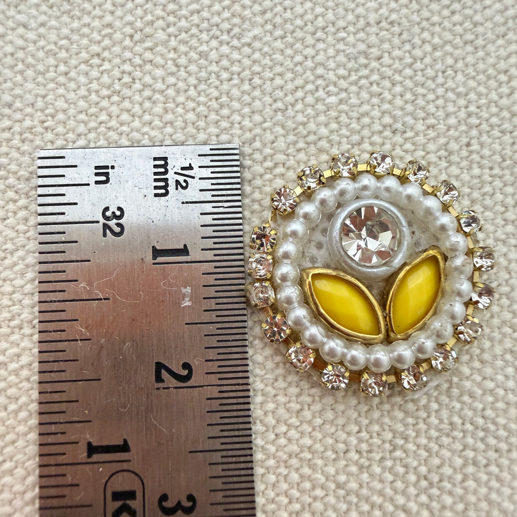 Decorative button with yellow center and pearl-like border next to a ruler for scale on a textured beige background