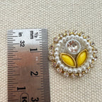 Decorative button with yellow center and pearl-like border next to a ruler for scale on a textured beige background