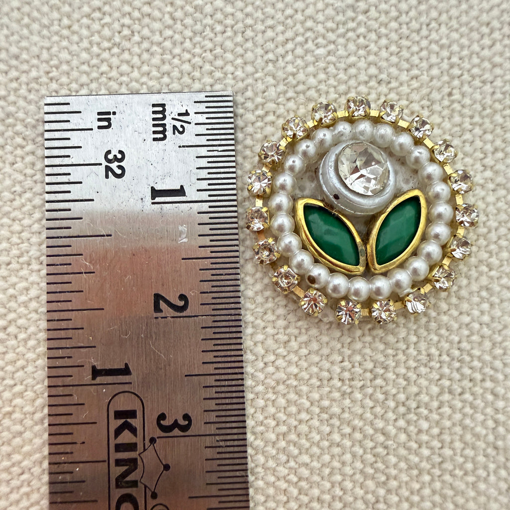 Decorative brooch with green leaves and pearl-like stones next to a ruler for scale on a textured beige background.