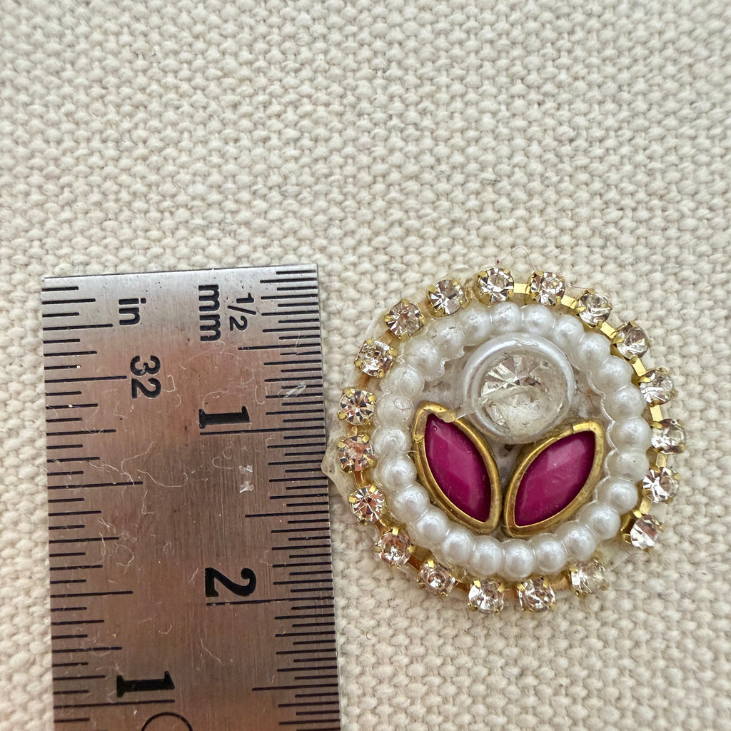 Decorative button with pink leaves and pearl details next to a ruler for scale on a textured beige background