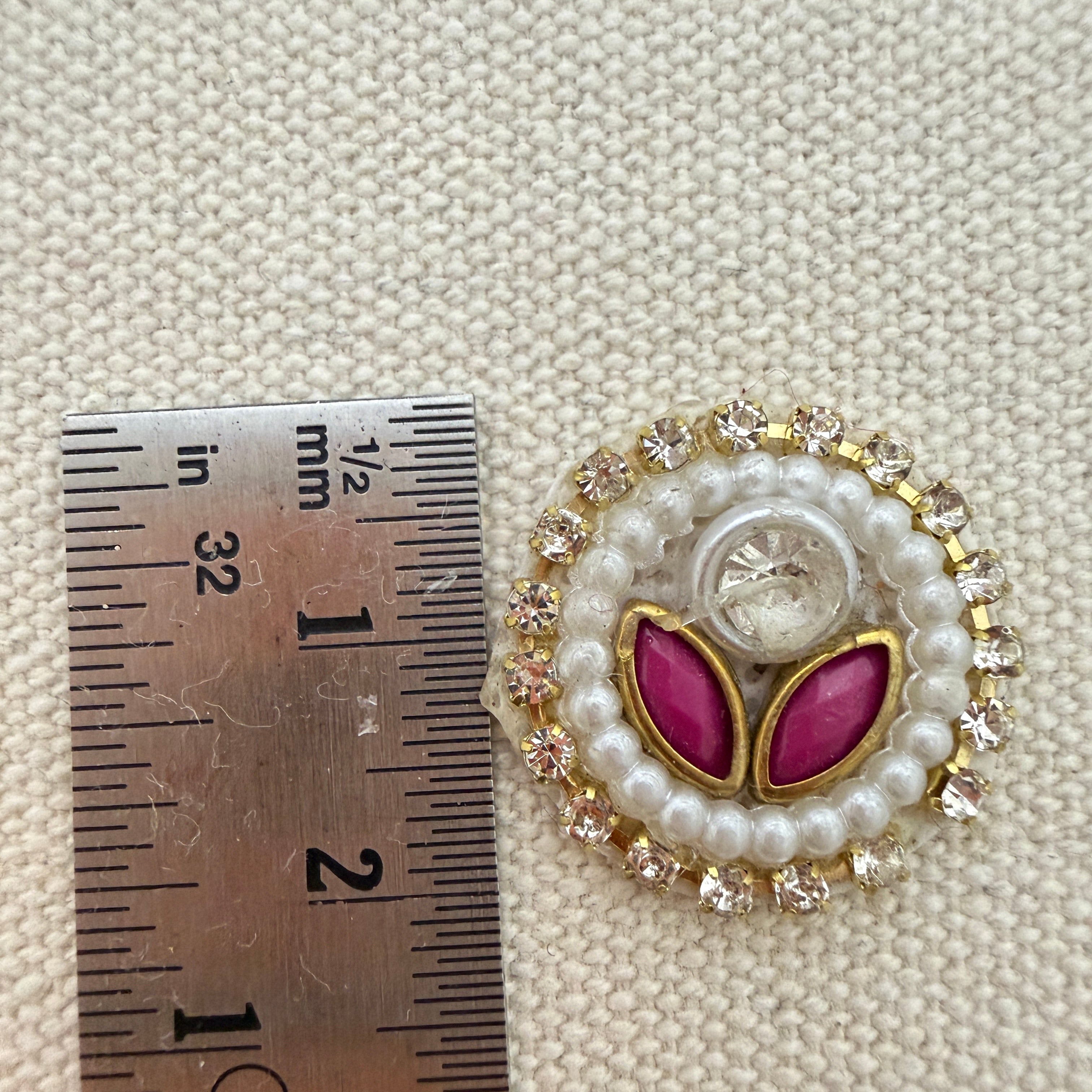 Decorative button with pink leaves and pearl details next to a ruler for scale on a textured beige background