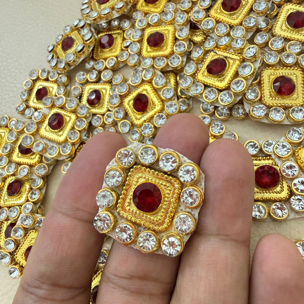 Hand holding ornate square gold-tone patch with ruby red center and clear crystal halo