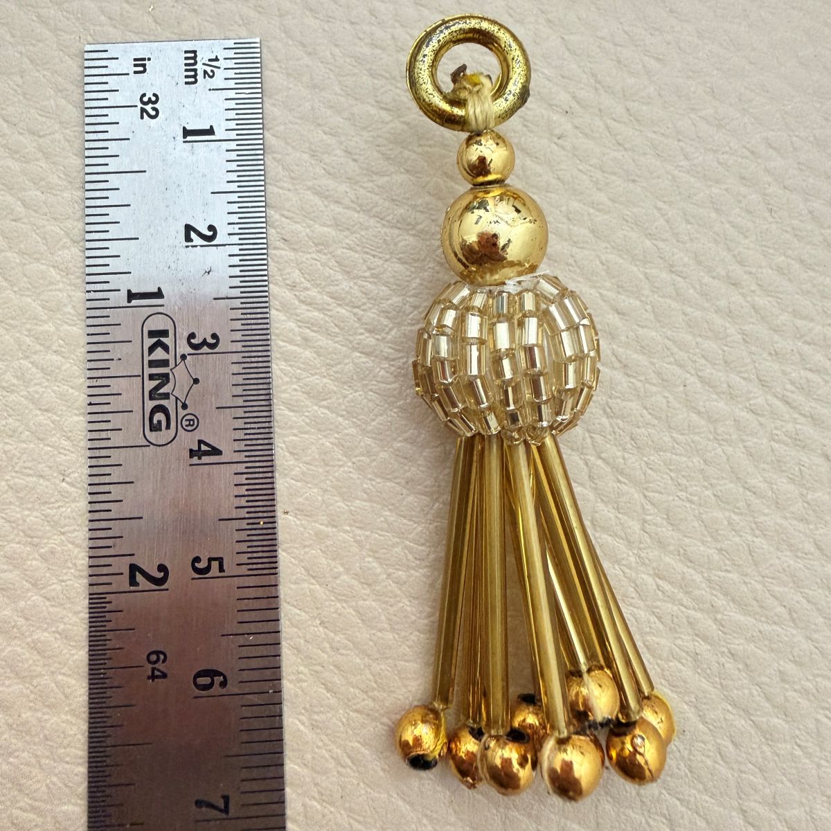 Gold metallic beaded tassel charm beside ruler measuring approximately 7 centimeters long