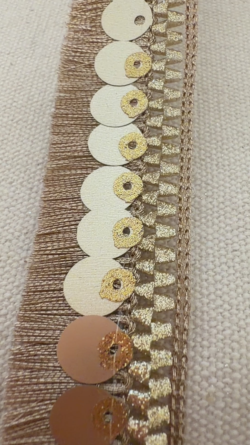 Close-up of a spool of decorative ribbon with gold sequins and fringe on a light background.