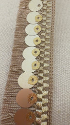 Close-up of a spool of decorative ribbon with gold sequins and fringe on a light background.