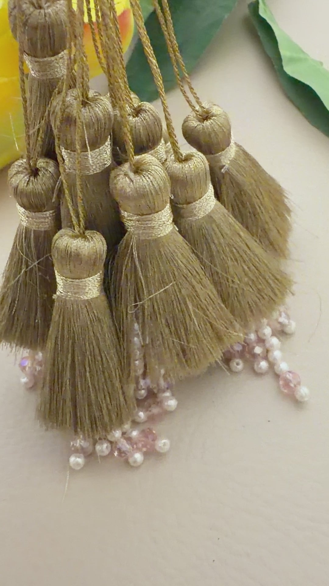 Gold tassels with pearl embellishments on a neutral background with yellow flowers.