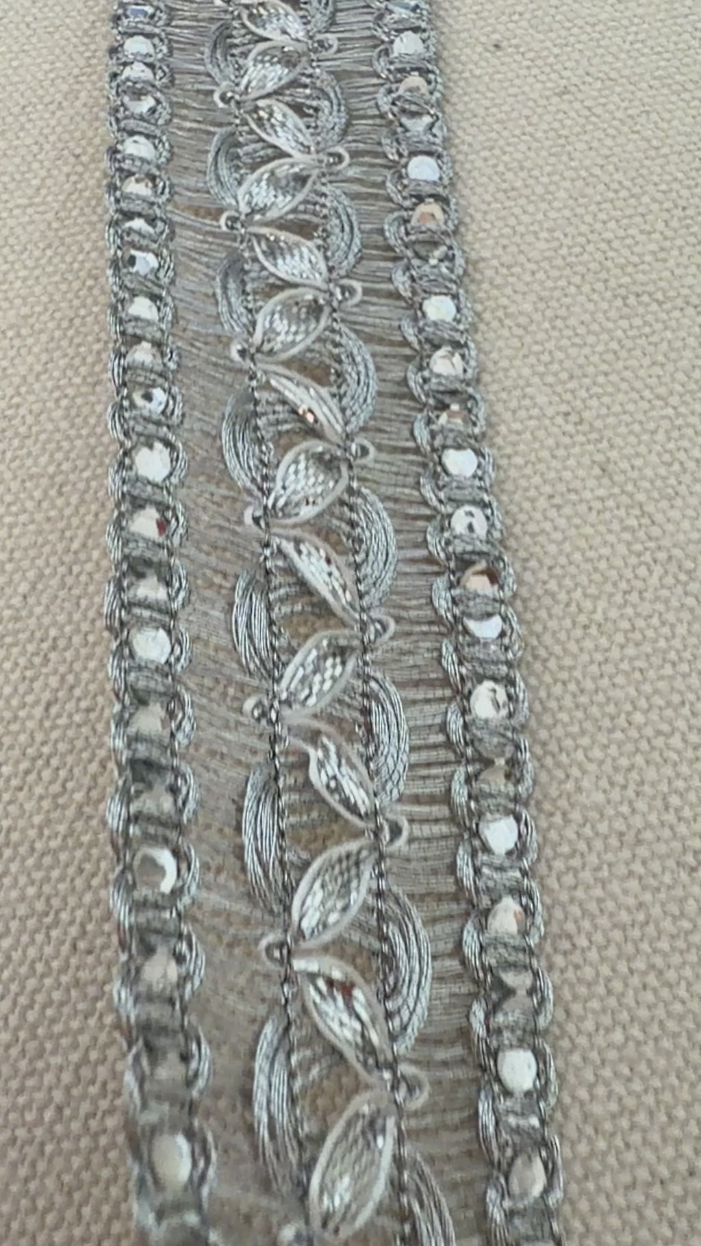 Silver decorative ribbon with intricate patterns on a beige background with a white flower.