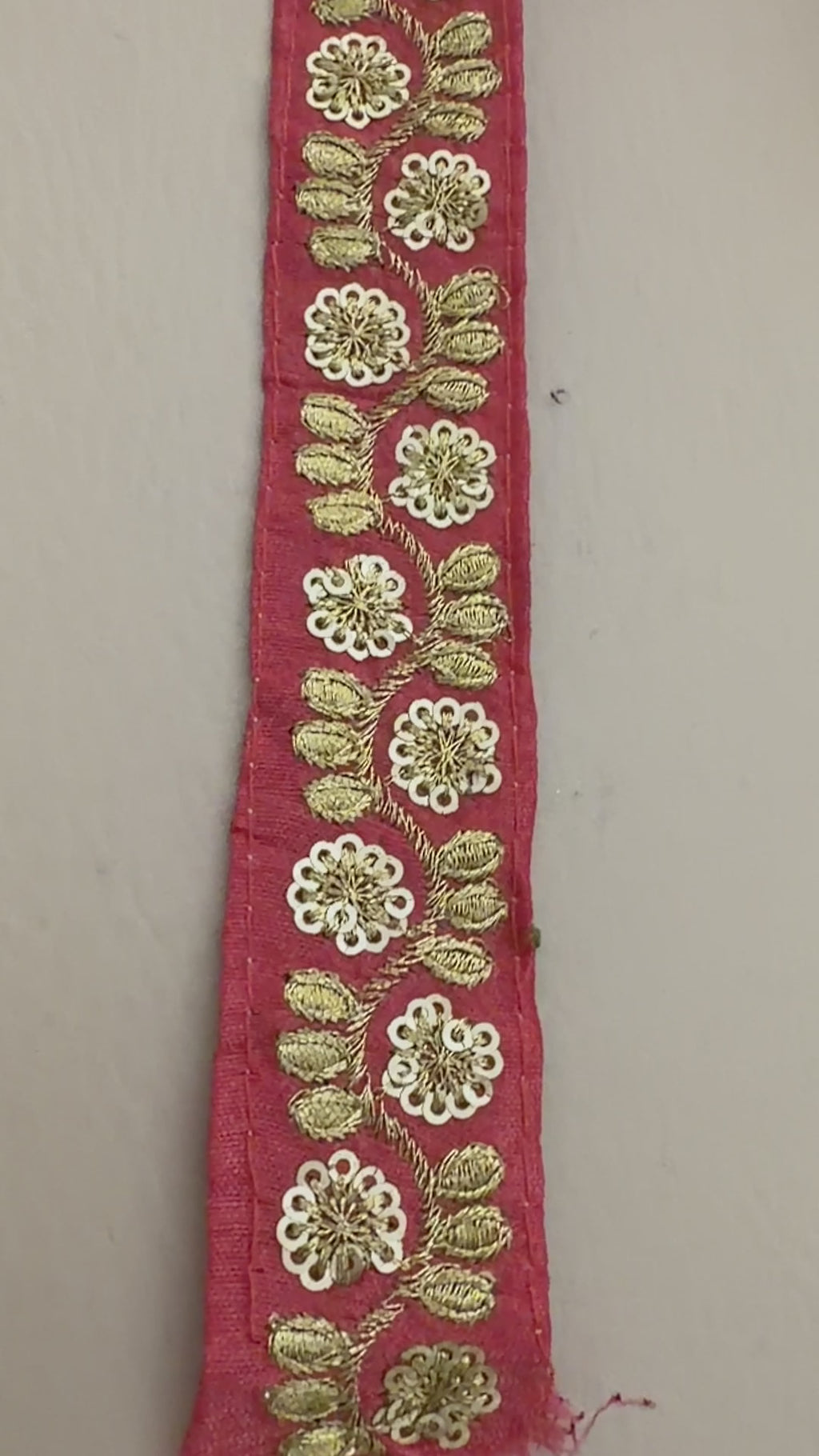 Red ribbon with gold floral embroidery held by a hand, with a roll of ribbon and flowers in the background.