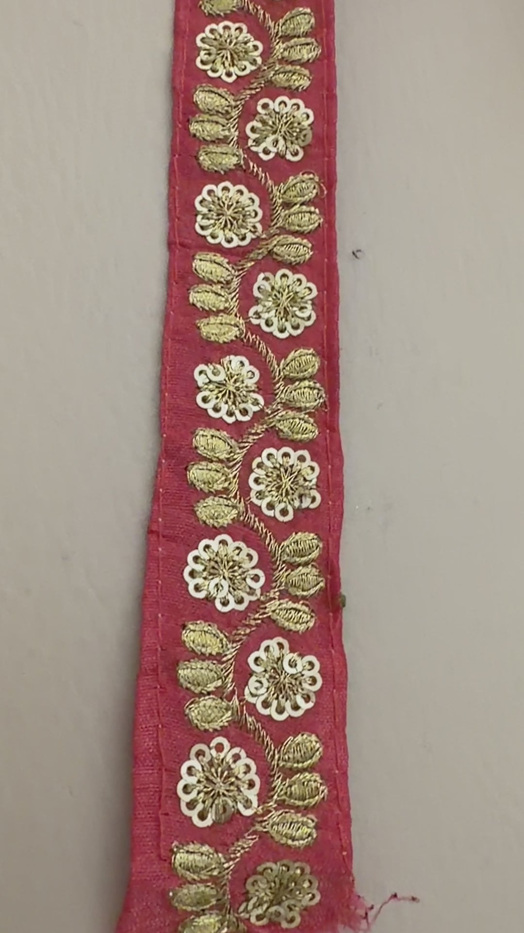 Red ribbon with gold floral embroidery held by a hand, with a roll of ribbon and flowers in the background.